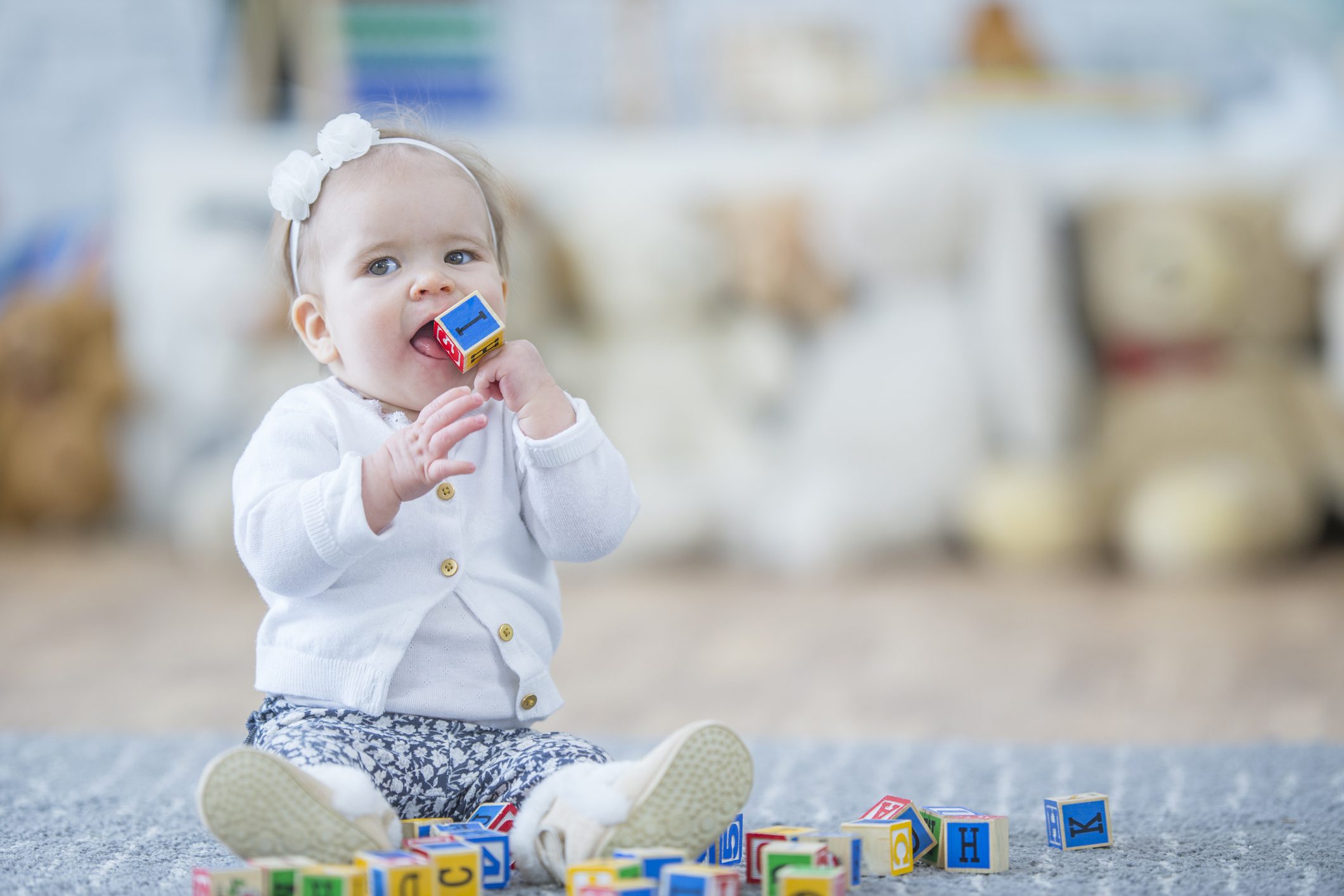 A baby girl is indoors in a daycare center. A baby girl is indoors in a daycare center. She is playing with toy blocks and trying to put one into her mouth.